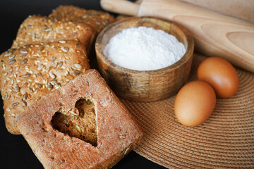 A cut out heart on a slice of bread next to small fresh breads with seeds and sesame on a wooden tray next to eggs, a wooden bowl with flour and a rolling pin