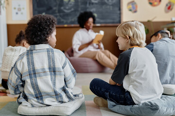 Back view of two younger children sitting on floor cushions in classroom listening to captivating story while looking at each other and secretly chatting