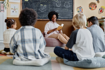Young female teacher of Black ethnicity in bean bag chair reading book together with children...