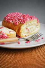 Sponge roll with cream and pink sprinkles on a white plate next to a fork on a gray background