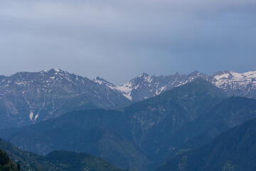 Majestic snow-capped mountains overlook a verdant valley under a cloudy sky, forming a stunning landscape. Peaks soar into the sky, framed by blue and white clouds