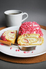 Sponge roll with cream and pink sprinkles on a white plate next to a fork and cup on a gray background