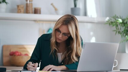 Video of pretty young business woman working with computer while consulting some invoices and documents in the kitchen at home. - Powered by Adobe