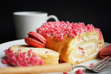 Sponge roll with cream, pink sprinkles and raspberry macaroons on a white plate next to a fork and cup on a dark background