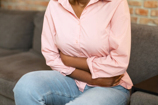 African American woman wearing a pink shirt and blue jeans sits on a couch with her hands clasped around her stomach. She appears to be in pain and discomfort, cropped