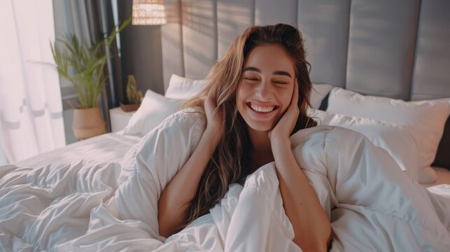 Beautiful young woman waking up in her bedroom, stretching hands with sleepy smiling face, sitting in bed under white linen duvet, had a good night sleep.