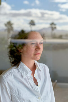 A thoughtful young woman with curly hair is gazing out a window, their reflection and distant trees mingling in the image.