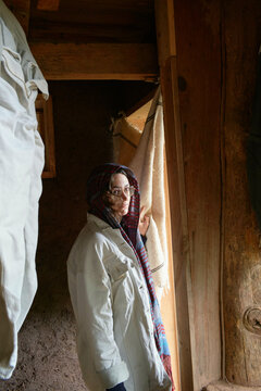 A young woman wearing a white coat and a colorful scarf stands by a wooden doorway, partially covered by a draped fabric, inside an apparent rustic building.