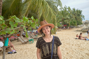 A smiling person wearing a brown hat stands on a sandy beach with greenery and sunbathers in the background.