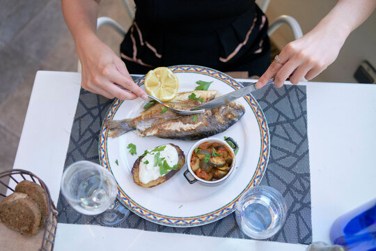 A person is seated at a table, about to enjoy a meal consisting of a grilled fish garnished with lemon, a side of bread, creamy sauce, and a small bowl of what looks like stewed vegetables.