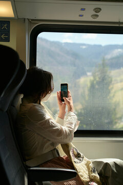 A person is seated inside a train, taking a photo of the mountainous landscape through the window with their smartphone.