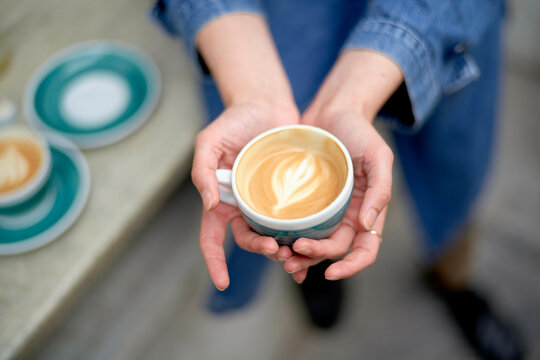 Hands cradling a ceramic cup filled with freshly brewed coffee, featuring a heart-shaped latte art, with more cups and saucers on a table in the blurred background.
