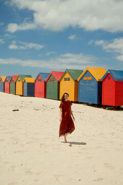 Woman in a red dress enjoying a sunny day at a beach with colorful beach huts in the background.