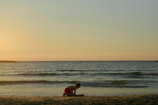 A child plays on a sandy beach at sunset with calm ocean waves in the background.