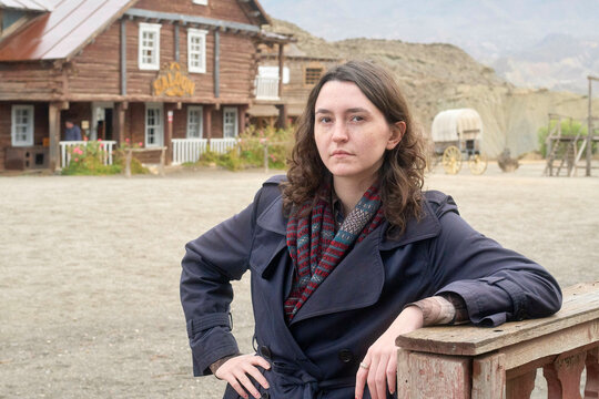 Confident woman leaning on a wooden fence in a western-style town setting with a thoughtful expression.