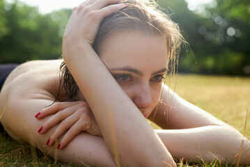Close-up of a thoughtful woman lying on the grass on a sunny day, with her head resting on her arms.