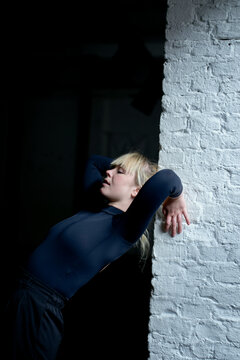 Woman in a dark blue top leaning against a white brick wall with a dramatic pose and shadow.