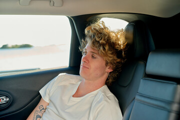 Curly-haired person relaxing in the backseat of a car with a pensive expression.