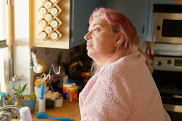 A woman with pink hair wrapped in a cozy beige towel stands in a kitchen with various items on the countertop.