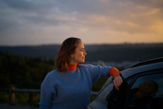 Woman leaning on her car enjoying a scenic sunset in the countryside.