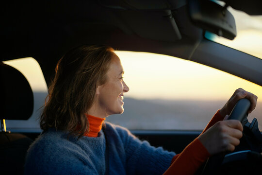 Smiling woman driving a car during sunset with warm light illuminating her face.