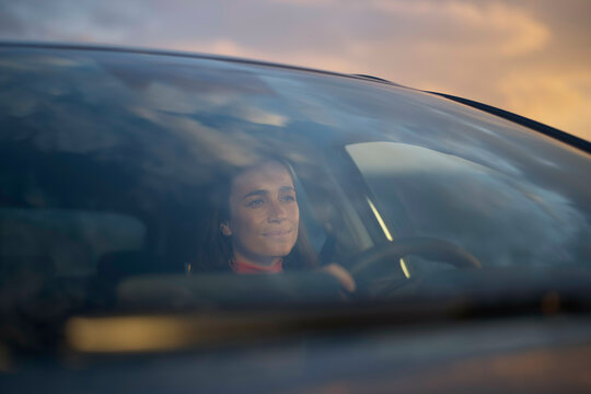 A smiling woman driving a car during sunset, with warm light reflecting on the vehicle's window.