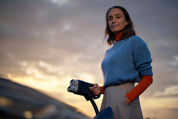 Confident woman holding an electric vehicle charging plug with a sunset sky in the background.