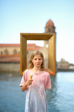 Smiling child holding an ice cream cone stands behind an empty picture frame with a historic building and water in the background.