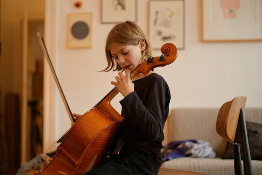 Young child practices playing cello in a cozy home environment with artwork on the wall.