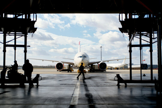 Silhouetted figures work around an aircraft inside a hangar, with clear skies in the background.