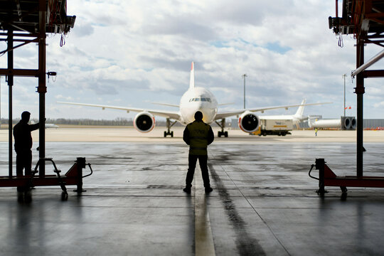 Ground crew member stands in an airplane hangar facing a parked commercial aircraft with another aircraft visible in the background.