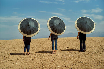 Three individuals stand side by side holding black umbrellas on a dry, sandy landscape under a cloudy sky.
