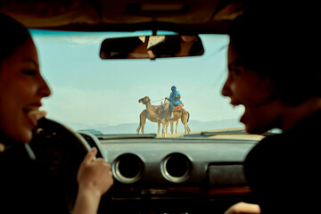View from inside a car where two individuals in the front seats are conversing and laughing, with a camel rider seen through the windshield in a desert landscape.