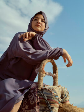 Woman in a purple hijab resting her arm on a rustic woven basket against a blue sky.