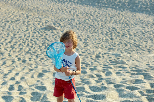 Young child holding a blue butterfly net on a sandy beach with sun casting long shadows.