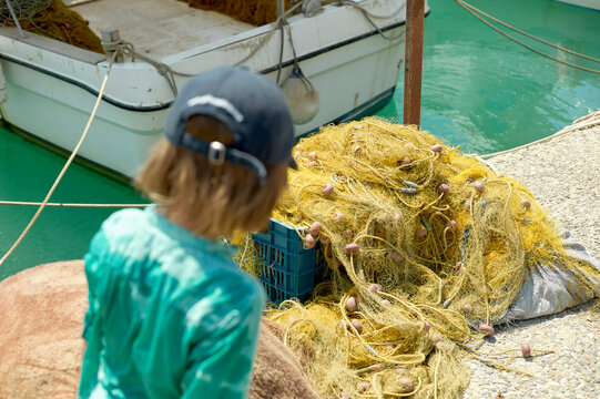 A child in a teal jacket observes a pile of yellow fishing nets beside boats moored on turquoise waters.