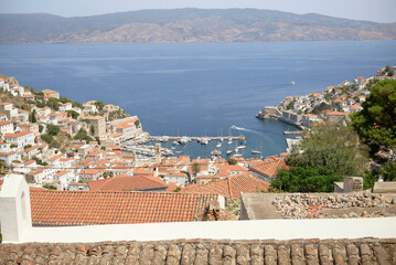Coastal town with terracotta rooftops overlooking a serene harbor with boats and clear blue waters, backed by distant hills.