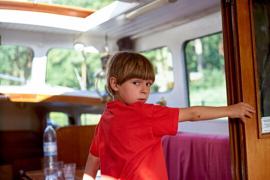 Young boy in a red shirt standing inside a bright camper van, turning to look at the camera with a thoughtful expression.