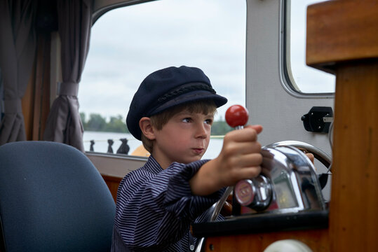 Young boy in a striped shirt and cap pretending to steer a boat with a focus on the wheel and throttle.