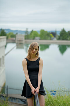 A young woman in a black dress stands by a railing with a serene lake and greenery in the background.