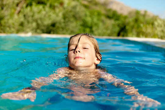 Child swimming in a pool with eyes closed and a calm expression during a sunny day.