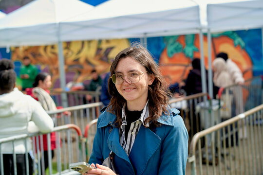 Smiling woman with glasses standing in front of white tents at an outdoor event with colorful murals in the background.