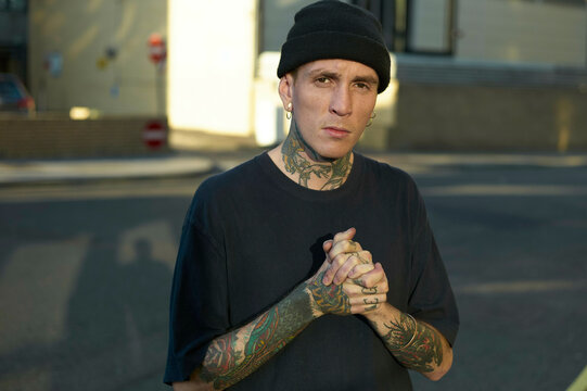 Tattooed man with a beanie posing with clasped hands on an urban street at sunset.
