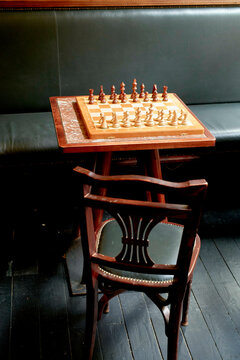 A chessboard with pieces neatly arranged for a game sits atop a wooden table next to a black leather couch, with an empty wooden chair facing it.