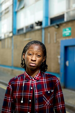 Person with dreadlocks wearing a red plaid shirt standing on an urban street with buildings in the background.