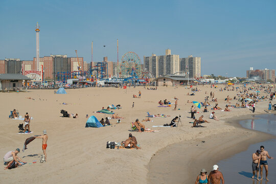 Sandy beach bustling with people on a sunny day, with an amusement park and city skyline in the background. - Powered by Adobe