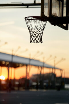 Sunset view through a basketball hoop at an outdoor court with a blurred cityscape in the background.