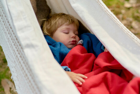A young child sleeps peacefully in a white hammock, wrapped in a cozy blue blanket.