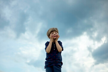 Young child standing against a cloudy sky, looking upwards with hands on cheeks in a gesture of surprise or awe.