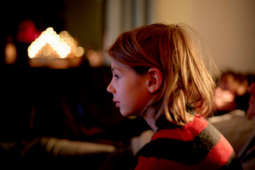 Side profile of a young girl focused on a glowing light source in a dimly lit room, with warm bokeh lights in the background.